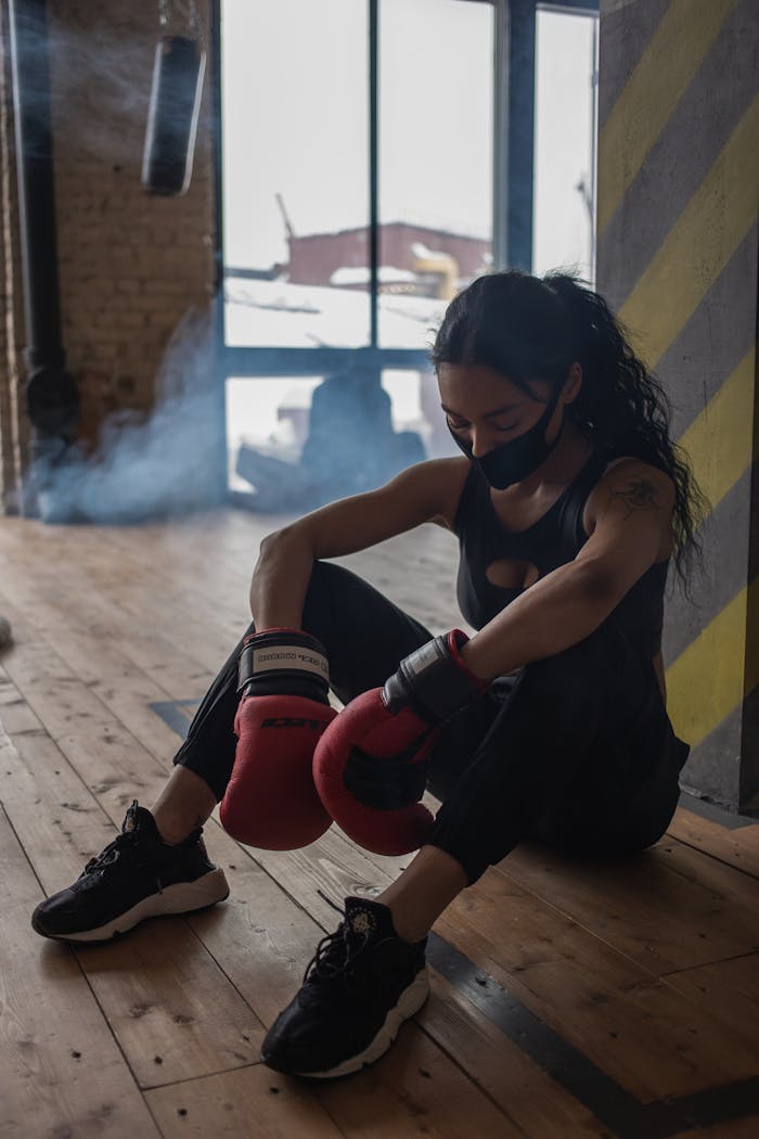 Home Exhausted black sportswoman in boxing gloves and cloth face mask having break from training on floor in gymnasium