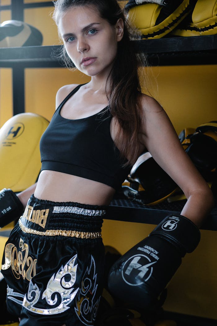 Portrait of a confident female boxer leaning in a gym, wearing black sportswear and boxing gloves.